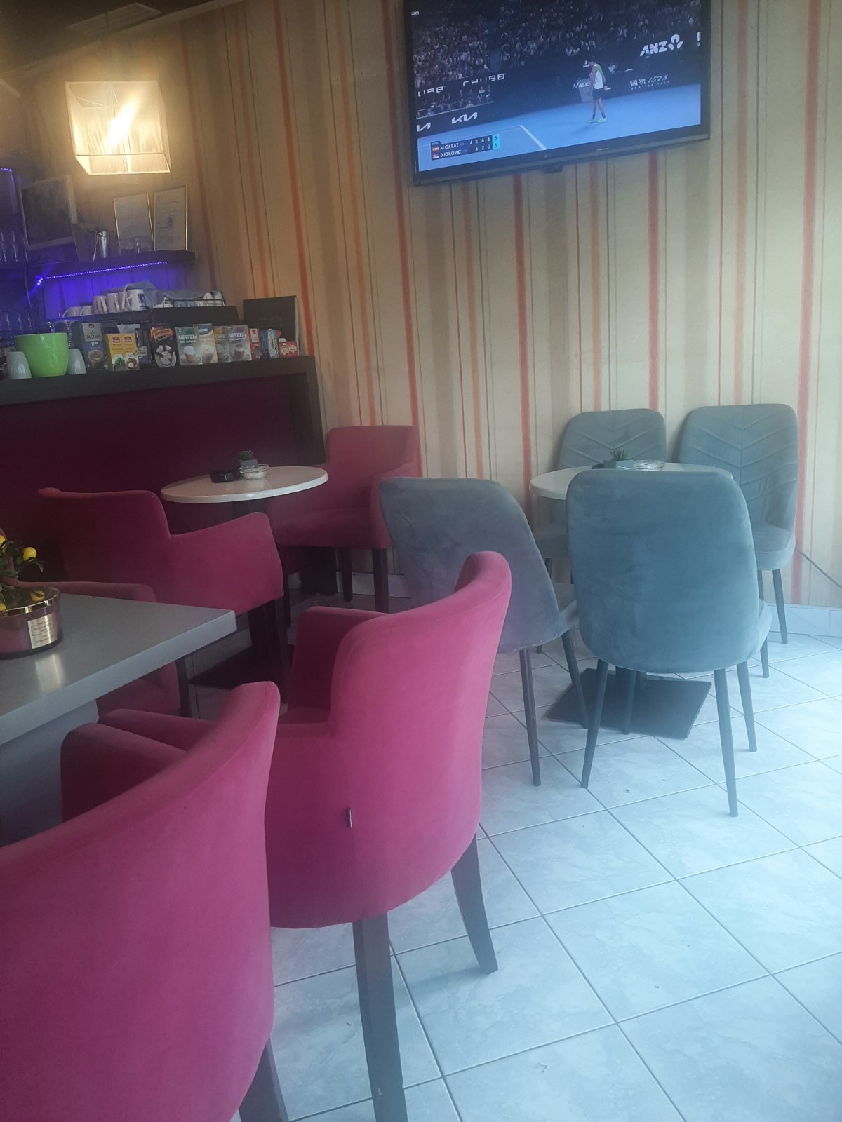 Fotelje boje ciklame i sive stolice, šank pod ljubičastim svjetlom i ekran u kafiću La Plaza